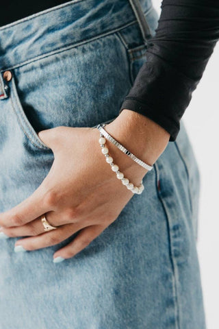 Person wearing blue jeans and a black long-sleeve shirt with a pearl bracelet on a neutral background