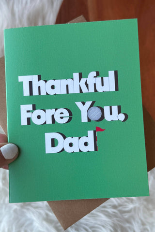 Green card with 'Thankful Fore You, Dad' text held by a hand on a soft surface.