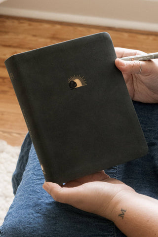 Person holding a black notebook with a logo on a wooden floor.
