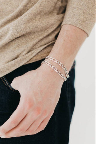 Person wearing a beige sweater and dark jeans with a silver bracelet on a plain background