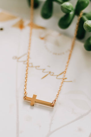 Gold necklace with a cross pendant on a marble surface with greenery in the background