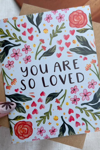 Greeting card with floral design and 'You are so loved' text held by a hand.