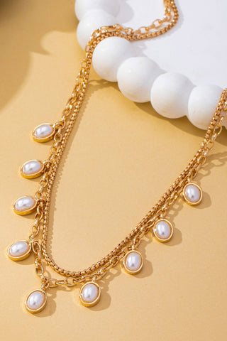 A gold-toned double-strand necklace with dangling oval pearl-like beads, styled on a warm beige surface with a white decorative plate in the background.
